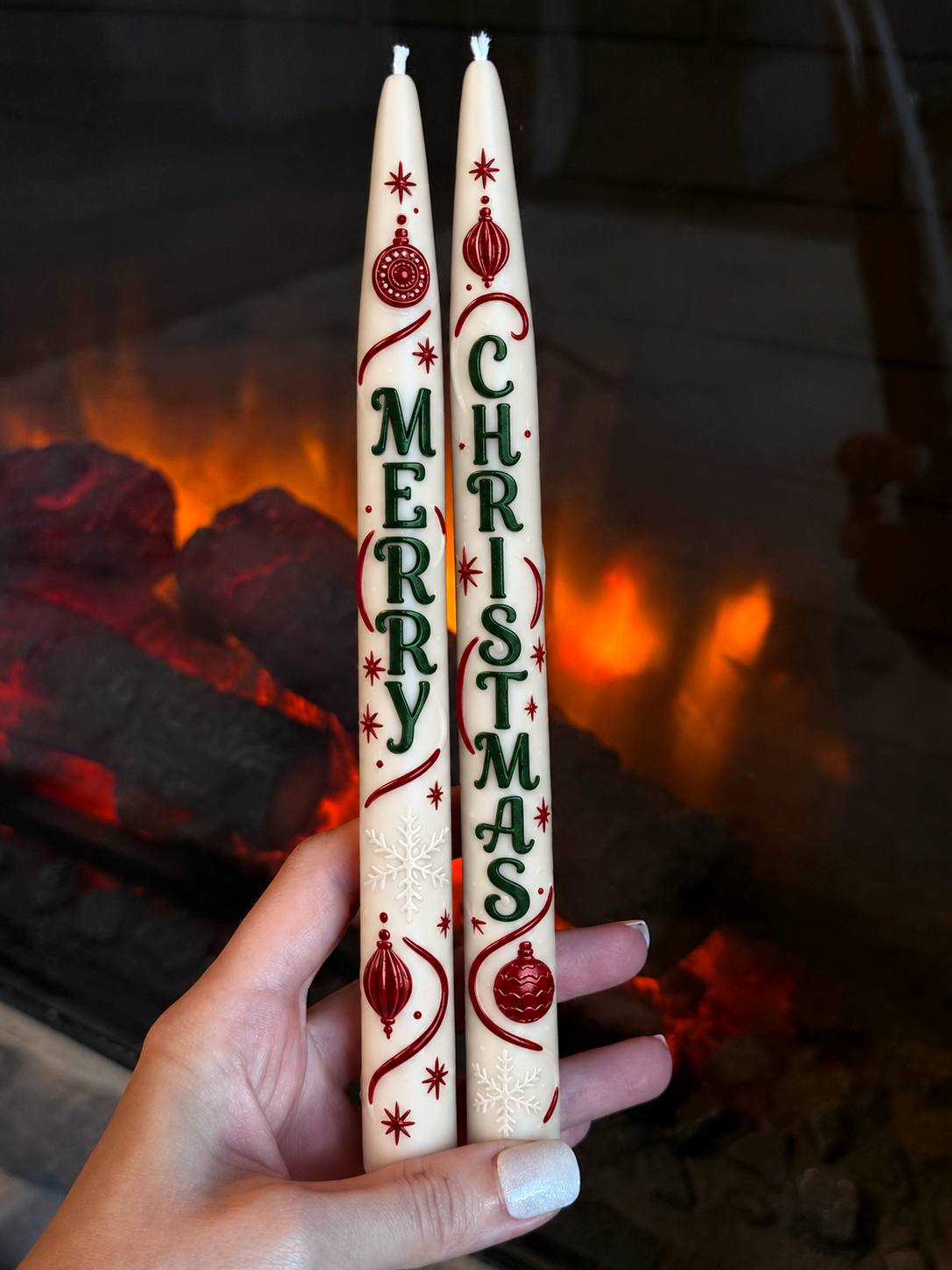 Two ivory Merry Christmas taper candles hand-painted in red and green and held in front of a glowing fireplace