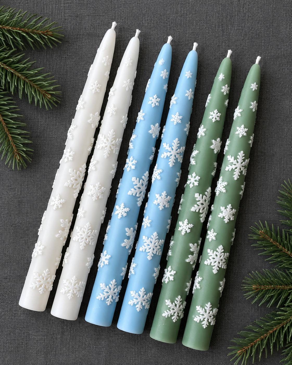 Six snowflake taper candles in white, light blue and sage green with raised white snowflakes, laid on grey linen with pine branches